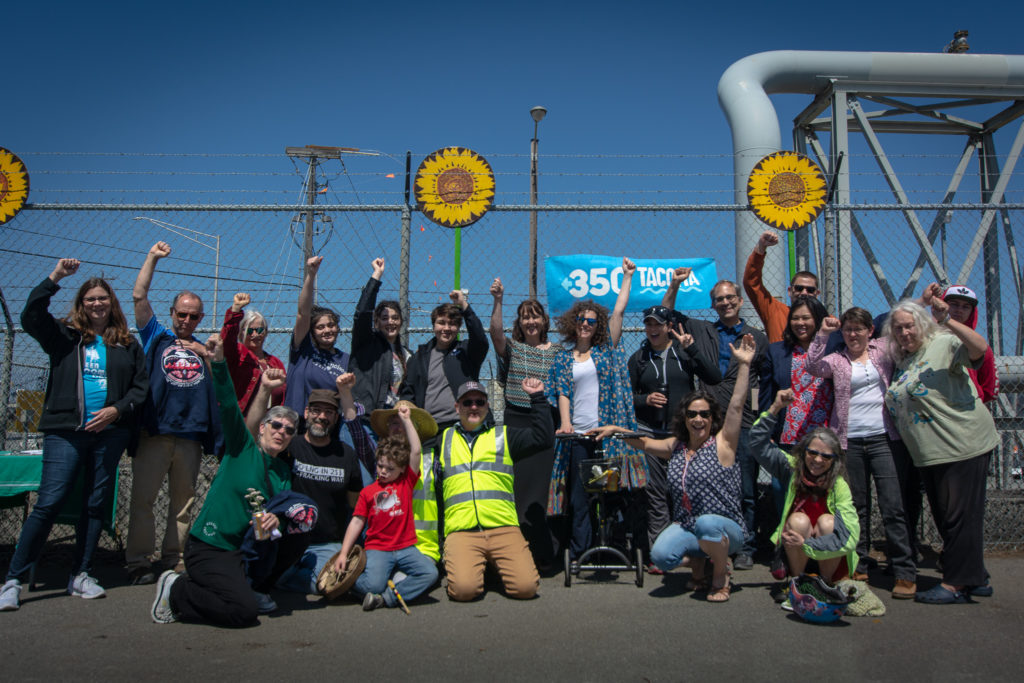 Group of 20 people smile and raise their fists in front of a chain-link fence with a "350 Tacoma" banner hanging